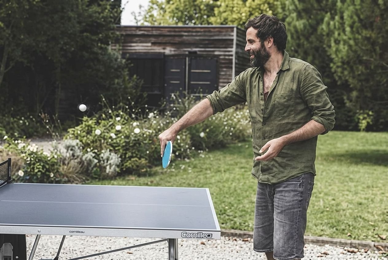 Homme jouant au ping-pong en extérieur, prêt à servir, table bleue et cadre verdoyant autour. table de ping pong exterieur ou interieur